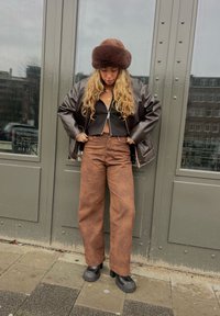 Brązowa skórzana kurtka pilotka, skrócony czarny top, brązowe spodnie o wysokim stanie i szerokich nogawkach oraz ciemne platformy; puszysty brązowy kapelusz.