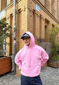 Lys rosa hettegenser med snor, frontlomme og vide ermer; kombinert med mørke jeans og en marineblå caps. Urban setting med mursteinsvegger.