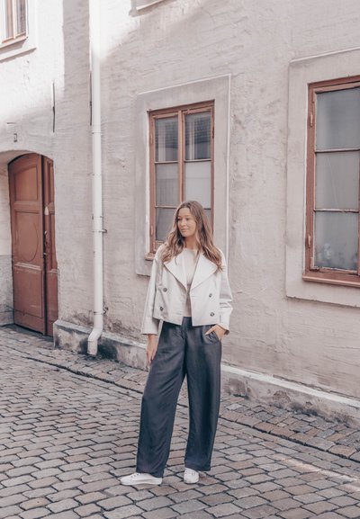 Chaqueta de doble botonadura de color claro, blusa crema, pantalones de pierna ancha grises, zapatillas blancas. Calle adoquinada y edificio beige de fondo.
