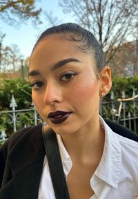 Persoon met donkere, glanzende lippenstift, een wit hemd en een oorsteker aan de oorschelp; haar naar achteren gebonden, lichte huid met subtiele sproeten, zacht licht.