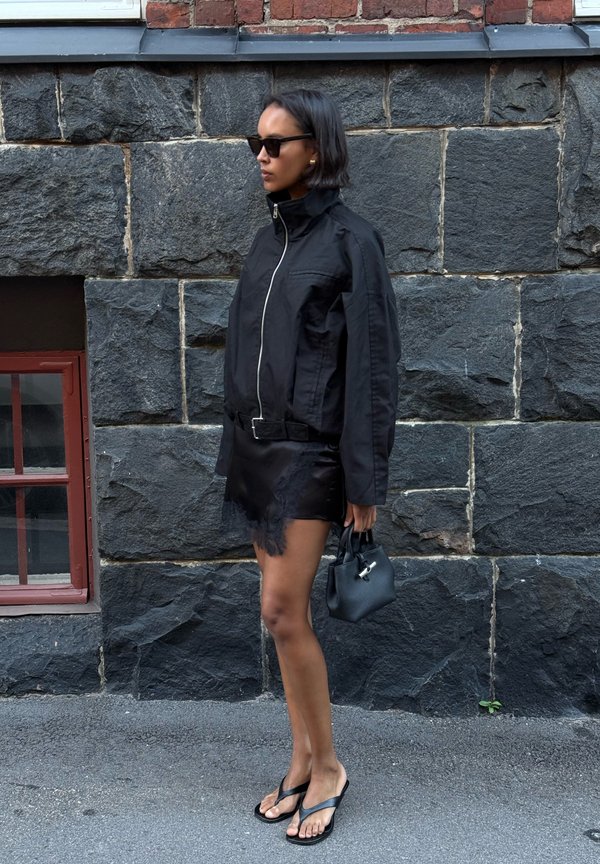 Mujer con pelo corto que lleva gafas de sol negras, chaqueta negra, falda corta, sandalias y sostiene un pequeño bolso negro, de pie junto a una pared de piedra.