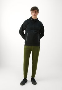 Sudadera negra con cremallera y bolsillos laterales, combinada con pantalones de chándal ajustados en verde olivo y zapatillas deportivas negras.
