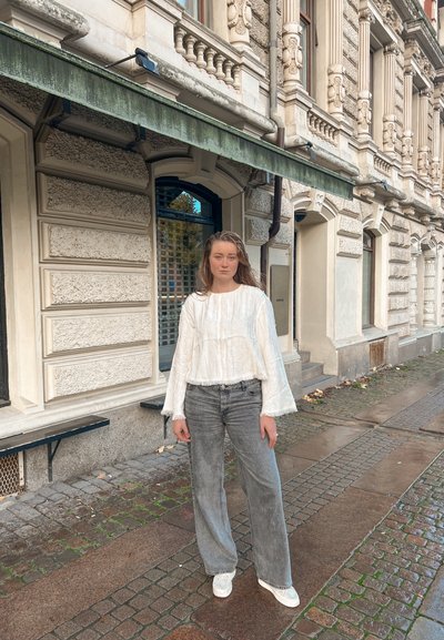 Joven con el cabello mojado de pie en una calle adoquinada mojada, vestida con una camiseta blanca de manga larga, jeans grises y zapatillas blancas, junto a un edificio ornamentado.