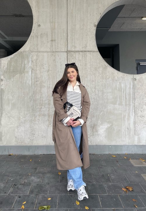 Černá kožená bunda, zeleno-modrý šátek, světle modré džíny, černé kotníkové boty. Stojí v parkovacím garáži s betonovými sloupy.