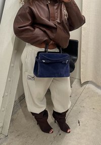 Personne portant une veste en cuir marron, un pantalon blanc avec un logo, et des bottines marron à talons, tenant un sac à main bleu marine à l'intérieur sur un sol en béton.