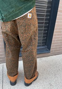 Cargo παντελόνια με σχέδιο λεοπάρ σε καφέ και μαύρο χρώμα, σε χαλαρή γραμμή; διαθέτουν πολλαπλές τσέπες και ραφτό λογότυπο Carhartt στο πίσω μέρος.