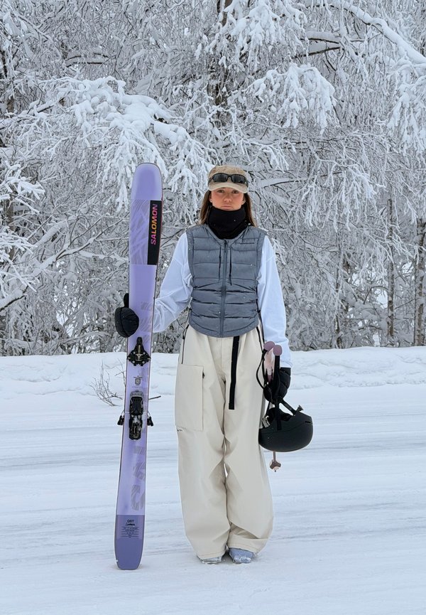 Oseba v zimski opremi stoji na snegu, v eni roki drži vijolični smučarski aparat Salomon, v drugi pa črno čelado, v ozadju pa so drevesa pokrita s snegom.