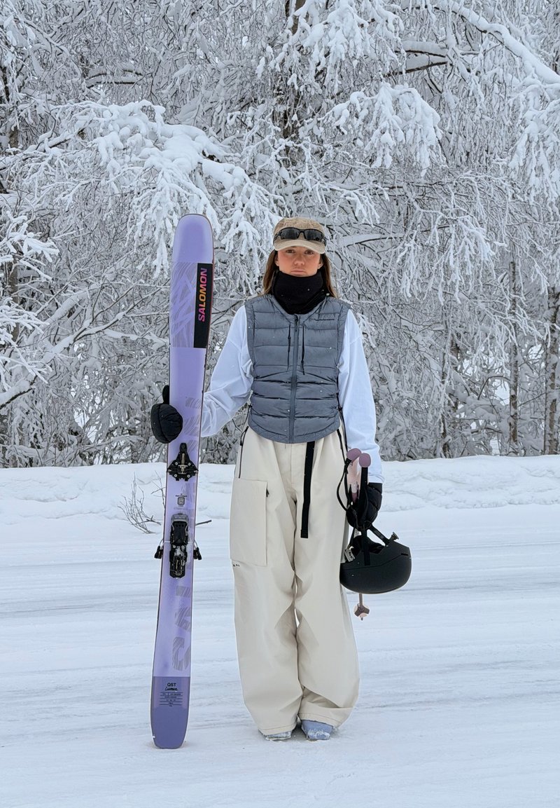 Άτομο με χειμερινό εξοπλισμό στέκεται στο χιόνι κρατώντας ένα μοβ σκι Salomon στο ένα χέρι και ένα μαύρο κράνος στο άλλο, με χιονισμένα δέντρα στο φόντο.