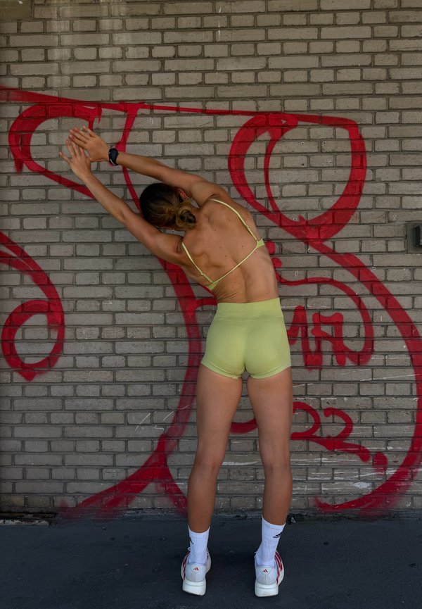Vrouw die zich uitstrekt met de armen boven het hoofd gestrekt tegen een met graffiti bedekte stenen muur, gekleed in lichtgroene korte broek en een sporttop.