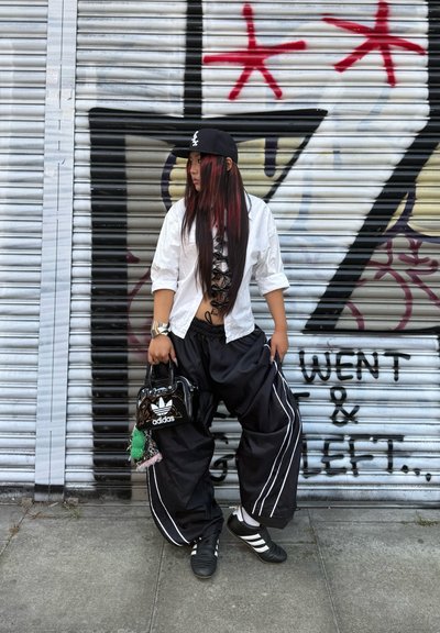 Pantalones oversize en blanco y negro con franjas blancas, blusa blanca con encaje, gorra negra, bolso de charol negro con el logo de Adidas y zapatos deportivos negros.