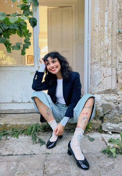 Zapatos de tacón de charol negro con punta cuadrada. Pantalones vaqueros de tiro corto y pierna ancha en azul claro, blusa blanca de manga larga y calcetines de encaje con un patrón floral.