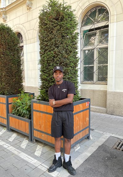Camiseta negra con un logo, pantalones cortos negros, botas de combate negras y calcetines blancos. De pie cerca de jardineras de madera con arbustos en un entorno urbano.