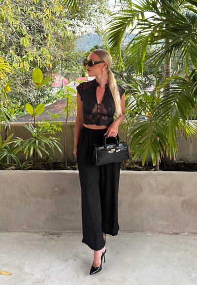 Mujer con pelo largo y rubio, con una blusa negra transparente y pantalones anchos, sostiene un bolso negro, de pie junto a una jardinera con plantas tropicales verdes.