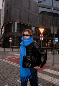 Veste en cuir noire, écharpe bleu vif et sac à main noir texturé. La personne se tient devant un bâtiment moderne dans un cadre urbain.