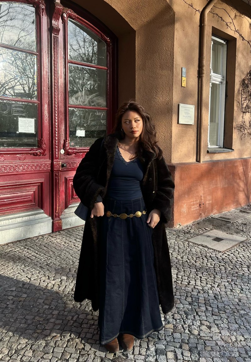 Femme en long manteau foncé, haut bleu marine et jupe, debout sur une rue pavée devant une porte rouge et un bâtiment beige avec une fenêtre.