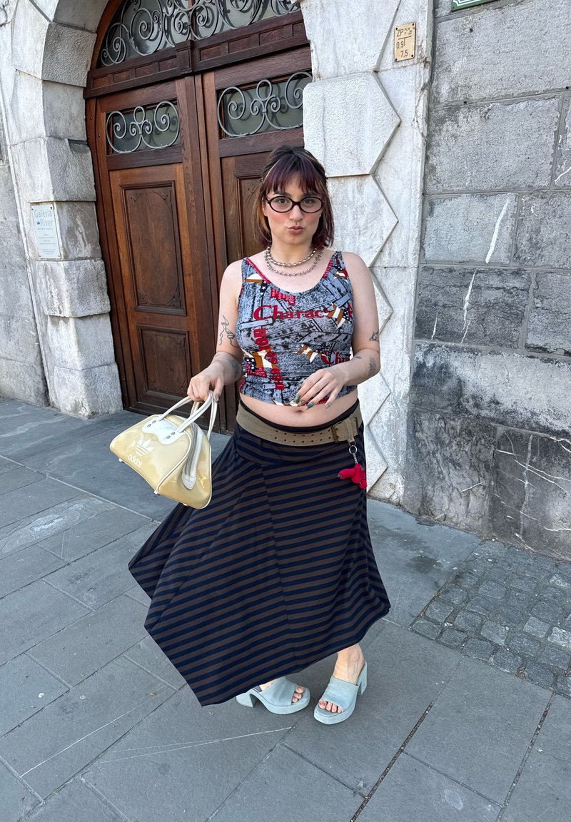 Jeune femme avec lunettes portant un crop top à motifs et une jupe rayée, tenant un sac beige, debout sur un trottoir en pierre près d'une porte en bois.