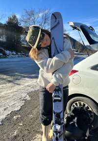 Skis Salomon au design blanc et violet, fixations noires ; portés avec un pull en polaire gris, un pantalon noir et des chaussures de ski crème. En arrière-plan, une voiture.