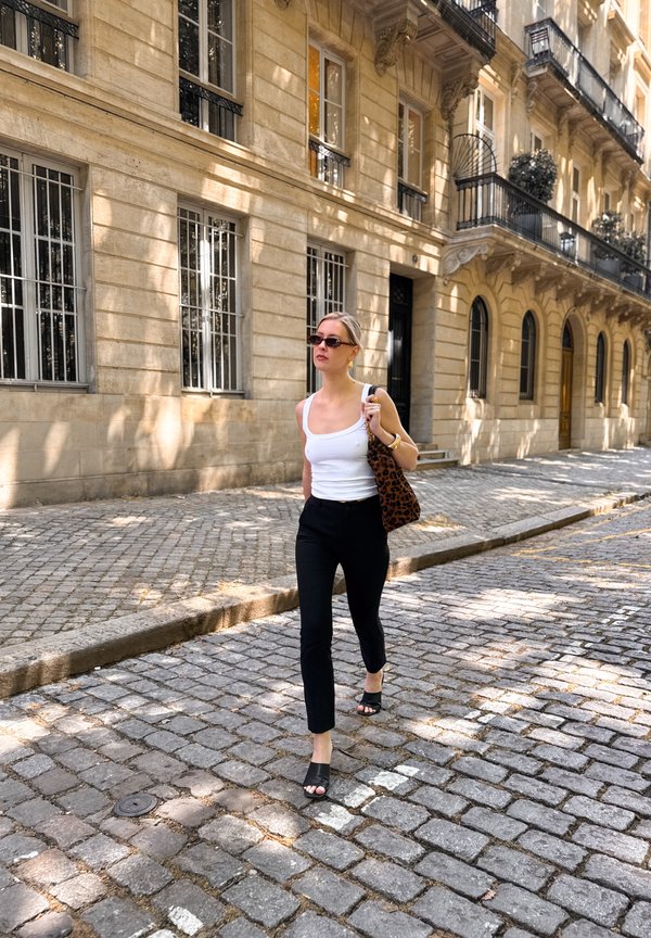 Mujer con camiseta sin mangas blanca y pantalones negros camina por una calle empedrada junto a un edificio de piedra beige con ventanas arqueadas y balcones.