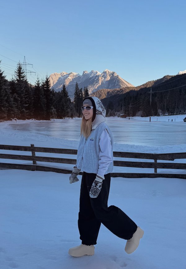 Jeune personne vêtue de vêtements d'hiver et de moufles marchant sur la neige avec en arrière-plan une montagne et une forêt de pins au coucher du soleil.