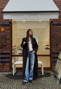 Dlhý hnedý semišový kabát cez károvanú košeľu a biele tričko, v kombinácii so širokými modrými džínsami a čiernymi topánkami, stojaci pred obchodom.