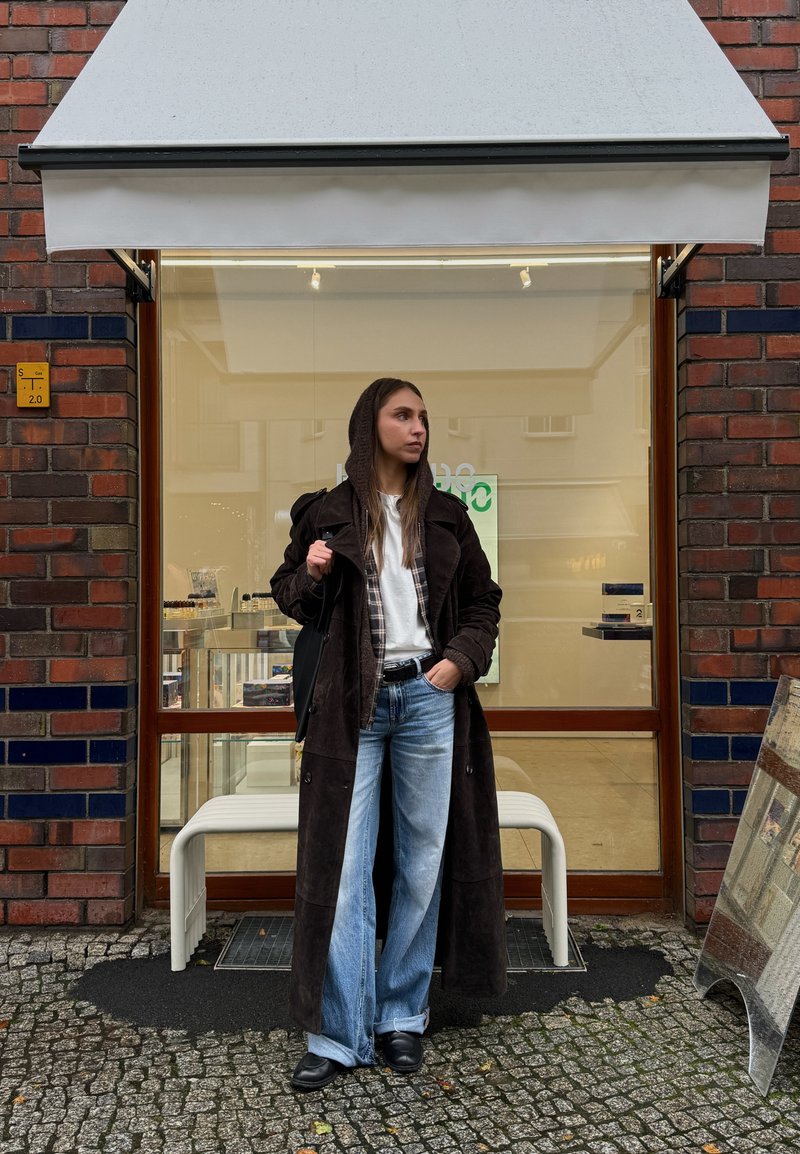 Casaco longo de camurça castanha sobre uma camisa xadrez e t-shirt branca, combinado com jeans azuis de perna larga e sapatos pretos, em frente a uma loja.