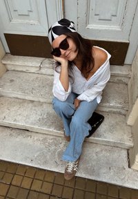 Modelo a usar uma camisa de linho branca, jeans claros de boca de sino e ténis castanhos, sentado em degraus de pedra perto de uma porta de madeira, com óculos de sol e um lenço estampado.