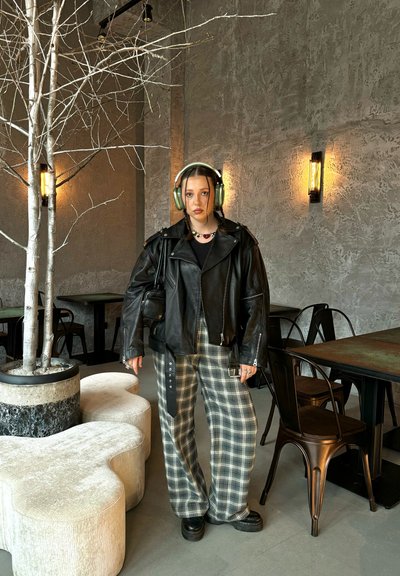 Chaqueta de cuero negra estilo motero, pantalones a rayas extragrandes en gris y blanco, zapatos negros gruesos y auriculares verdes. El entorno presenta paredes texturizadas.