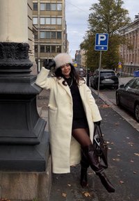 Cappotto di lana color crema sopra un vestito nero, guanti neri e stivali marroni alti fino al ginocchio. Tiene una borsa marrone, in piedi vicino a un lampione.