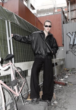 Chaqueta de cuero negra, pantalones negros sueltos, gafas de sol, apoyado en una valla; una bicicleta rosa cerca; entorno urbano con graffiti y escombros.