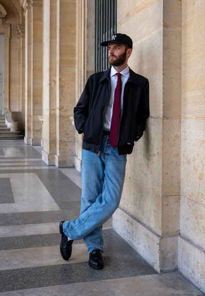 Hombre con barba, gorra, chaqueta negra, corbata roja y vaqueros apoyado contra un pilar de piedra en un pasillo con arcos.