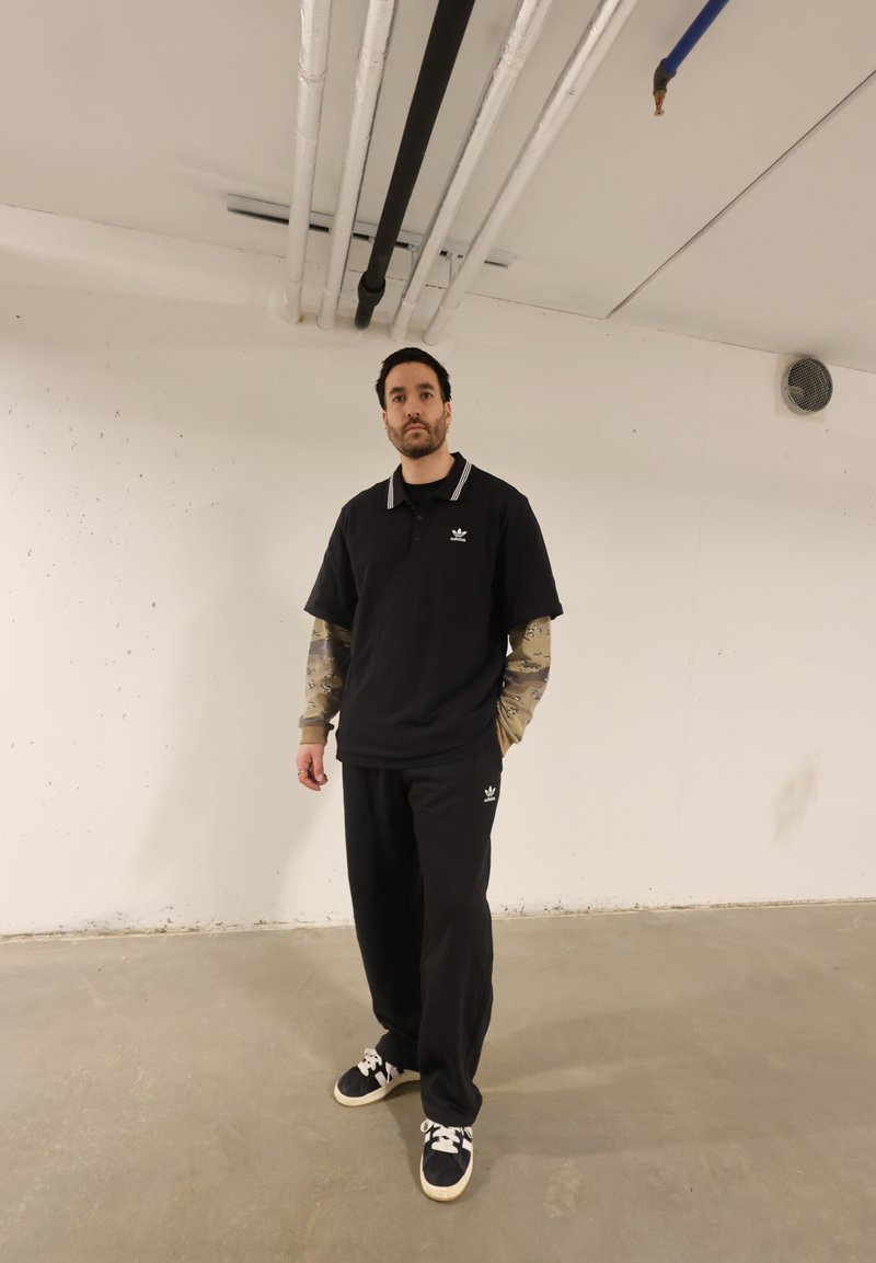 Homme debout dans une pièce simple, portant une tenue sportive Adidas noire avec des manches camouflage et des baskets noires à lacets blancs.