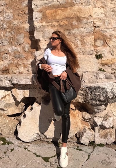 Mujer con cabello largo que lleva gafas de sol, camiseta blanca, pantalones negros y zapatos blancos, apoyada contra una pared de piedra con textura a la luz del sol.