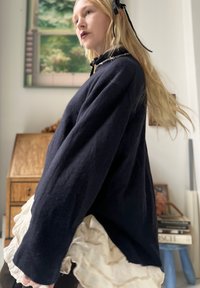Mulher loira com um laço preto no cabelo, vestindo um suéter escuro oversized e uma saia branca em camadas, de pé dentro de casa perto de móveis de madeira.