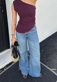 Burgundowy top na jedno ramię, marszczony, w połączeniu z jasnoniebieskimi spodniami typu wide-leg. Dodatki obejmują czarną torbę i słomkową torebkę.