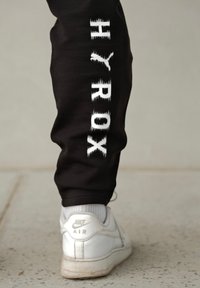 Czarne dresy z pionowym napisem "HYROX" w postarzonym białym kolorze. Połączone z białymi sneakersami Nike Air z delikatnym szarym akcentem.
