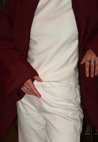 Burgundská prošívaná bunda přes bílý strukturovaný top a bílé proužkované kalhoty. Na prstech jsou dva stříbrné prsteny, ruce jsou umístěny v blízkosti pasu.