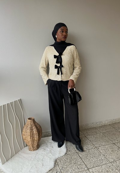 Cárdigan de textura crema con acentos de lazo negro, combinado con pantalones anchos negros. Un bolso negro y un hijab complementan el conjunto.