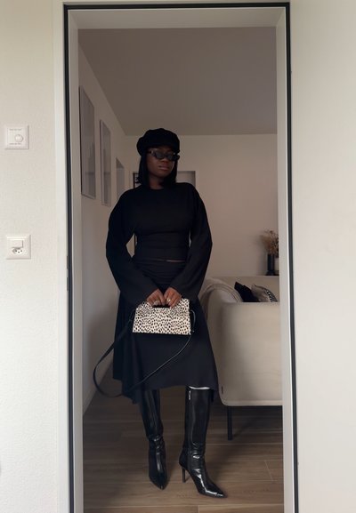 Mujer con vestido negro, boina negra, gafas de sol y botas negras hasta la rodilla, sosteniendo un pequeño bolso blanco con manchas negras en el interior.