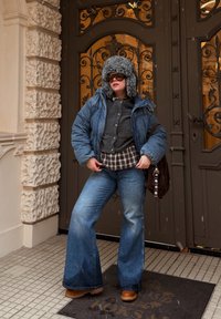 Riflová péřová bunda s kapucí lemovanou umělým kožíškem, vrstvená přes šedý svetr a károvanou košili. Pohled doplňují rozšířené modré džíny a hnědé boty.