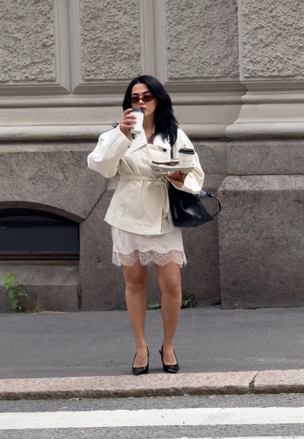 Femme en manteau blanc et jupe tenant deux tasses de café et une assiette, debout sur un trottoir devant un mur en pierre texturé.