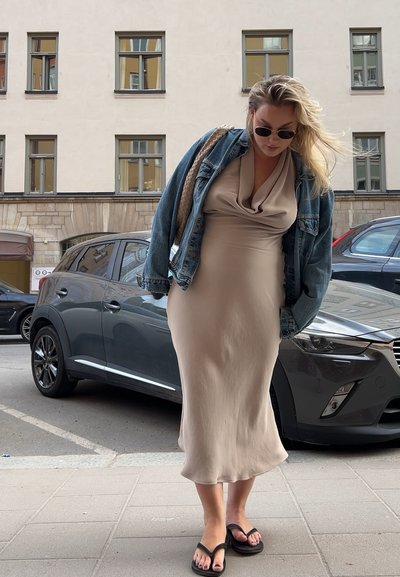 Vestido beige hasta la rodilla con un escote drapeado, combinado con una chaqueta de mezclilla azul. Chanclas negras. Coche gris aparcado al fondo.