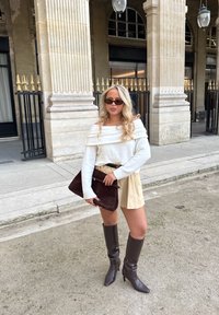 Offwhite off-shoulder genser, beige miniskjørt med frynset kant, brun semsket skinnclutch og brun skinnstøvel med hæl som går til kneet.