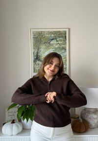 Ciemnobrązowy żeberkowany sweter z kołnierzem na suwak. Białe spodnie. Wnętrze z rośliną doniczkową, dekoracyjną dynią i oprawionym dziełem sztuki.