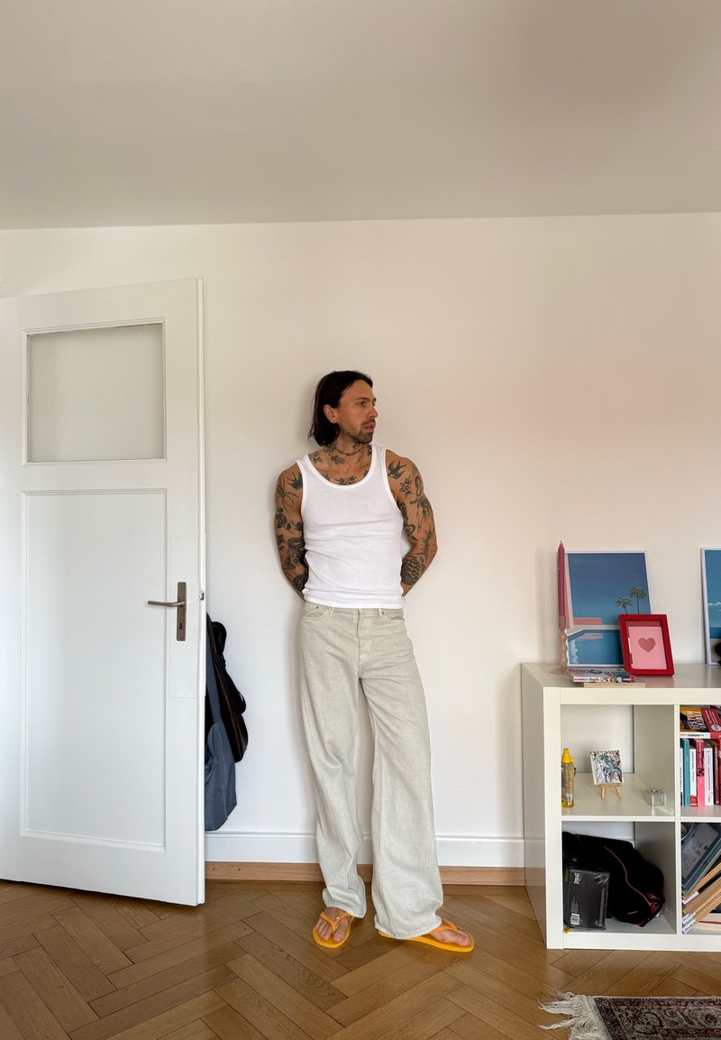 Homme tatoué en débardeur blanc, pantalon beige et sandales orange, debout près d'une porte ouverte à côté d'une étagère blanche avec des livres et des photos encadrées.