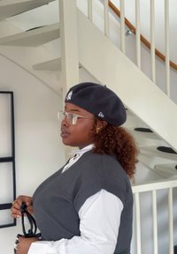 Szürke kötött mellény, fehér galléros ing, fekete logós barett, tiszta szemüveg, göndör haj, egy kis fekete táskát tart. Fehér lépcső van a háttérben.