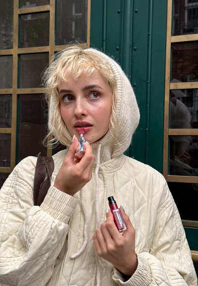 Modelo con una chaqueta de crema acolchada y capucha de punto blanca aplicándose brillo labial. Sostiene un producto labial transparente con un tinte rojo y tapa negra. Fondo verde.