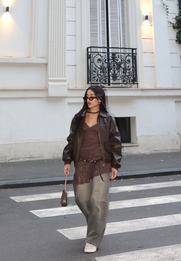 Bruine leren jas boven een bordeauxrode kanten top, gecombineerd met losse grijze jeans. Accessoires zijn zwarte zonnebrillen en een kleine bruine tas.