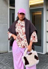 Přes oversized hnědé svrchní oblečení je přes oblečený vzorovaný top; růžové pruhované široké kalhoty; chlupatá bílá kabelka s hnědými akcenty; růžová čepice.