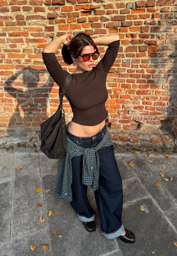 Una mujer posa junto a una pared de ladrillo con gafas de sol naranjas, una camiseta oscura, vaqueros holgados y una camisa de cuadros atada a la cintura. Lleva un bolso negro.