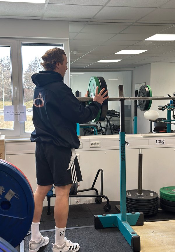 Sollevatore di pesi che regola i pesi su un bilanciere, indossando un felpa nera e pantaloni corti, con attrezzatura di un mix di verde, nero e blu in una palestra.